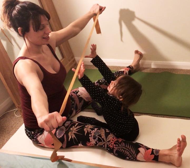 Kat Frost, Yoga2shape, sits in upavista konasana (wide angle pose) holding a ribbon for the two year old toddler to grab.