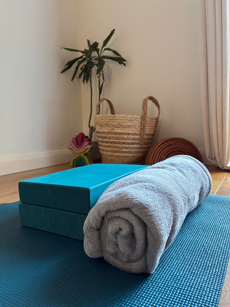 Calm and comfortable yoga studio with a mat, rolled blanket and yoga blocks plus decorative items such as earthy wooden ornaments, basket and plant.
